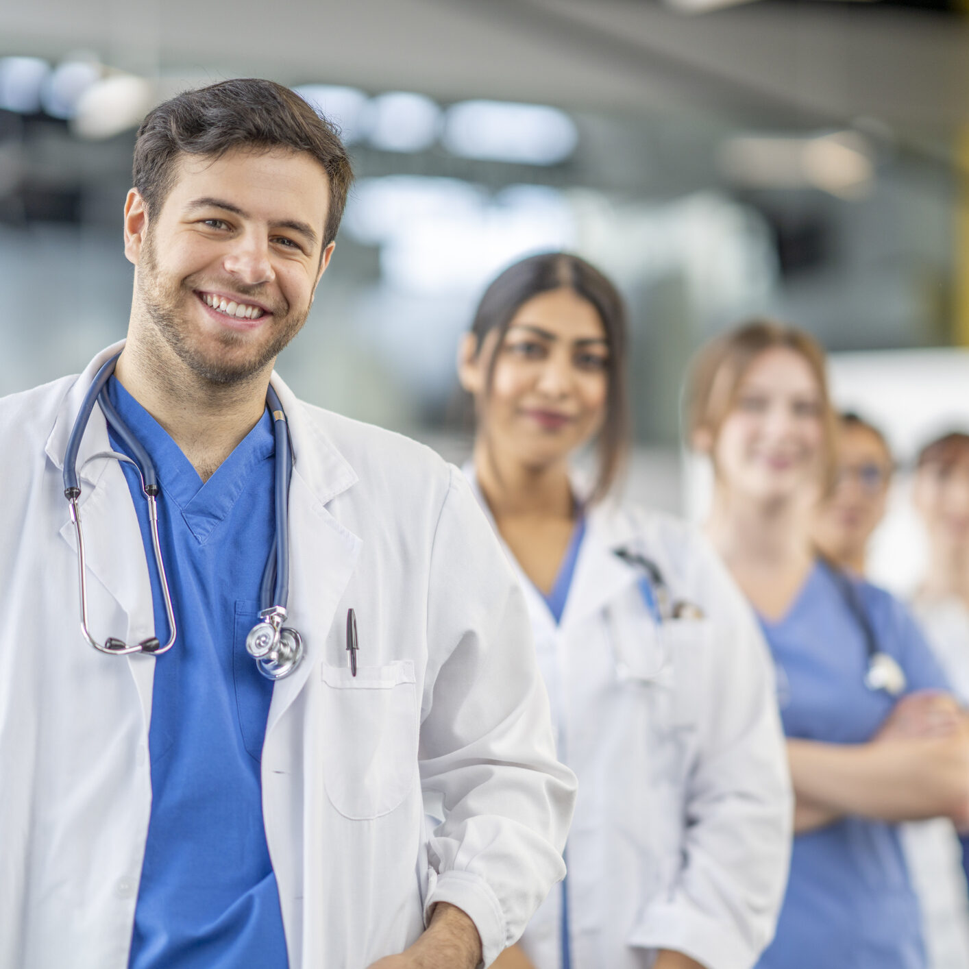 A small medical team stands in the hallway of a hospital together as they pose for a portrait.  They are each dressed professionally in scrubs and lab coats as they smile.  The focus is on a male doctor in front who is leading the team.